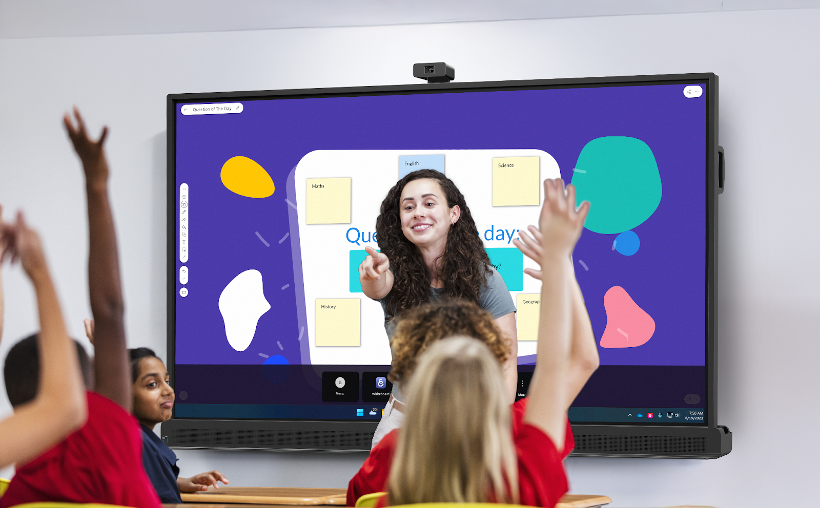  A teacher stands at the front of her classroom, pointing toward students raising their hands. Behind her, an interactive display shows the “Question of the Day.”