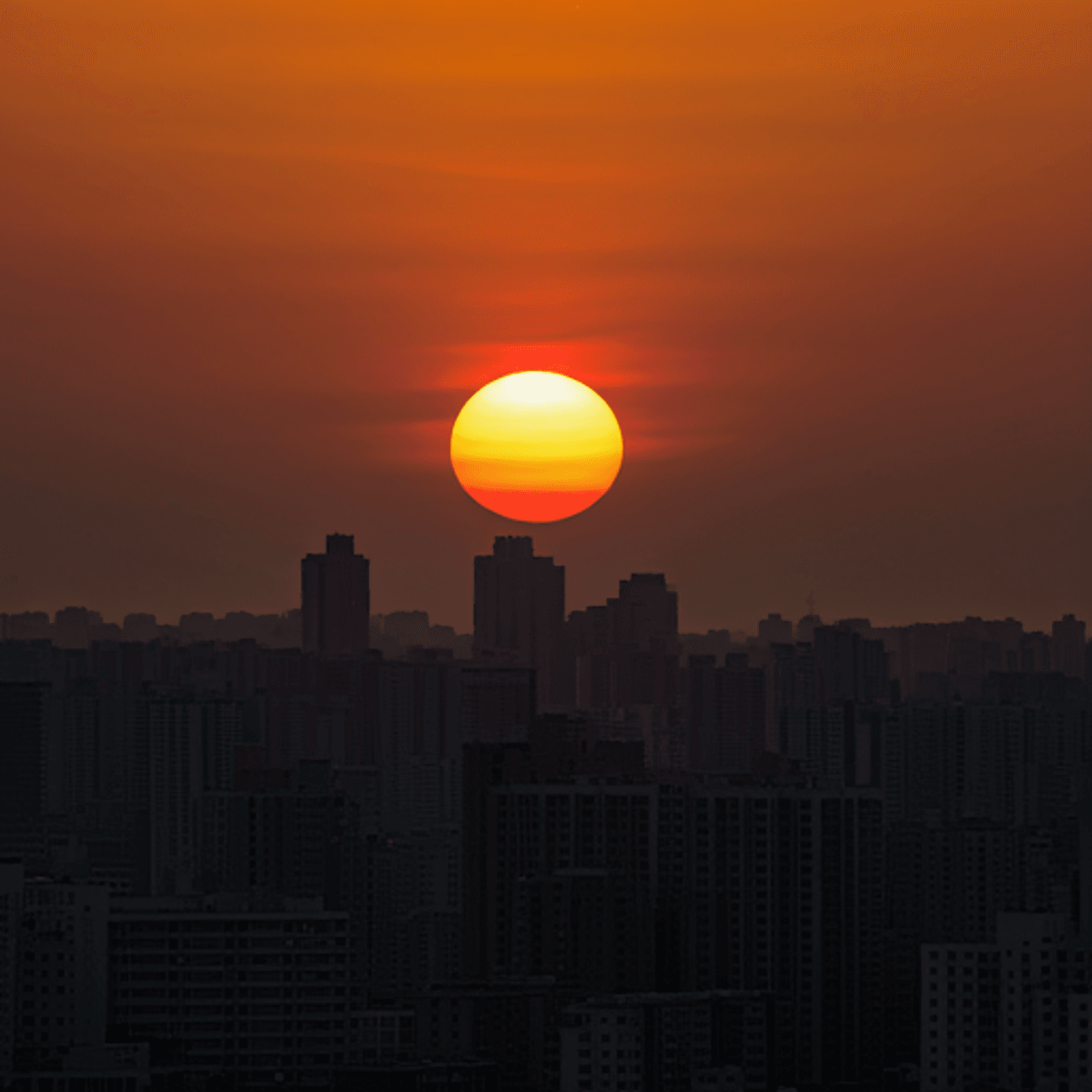 A vibrant sunset with a glowing orange sun setting behind urban city skyscrapers.