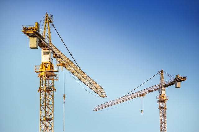 Two large yellow construction cranes tower against a clear blue sky, their long horizontal arms poised over the site.