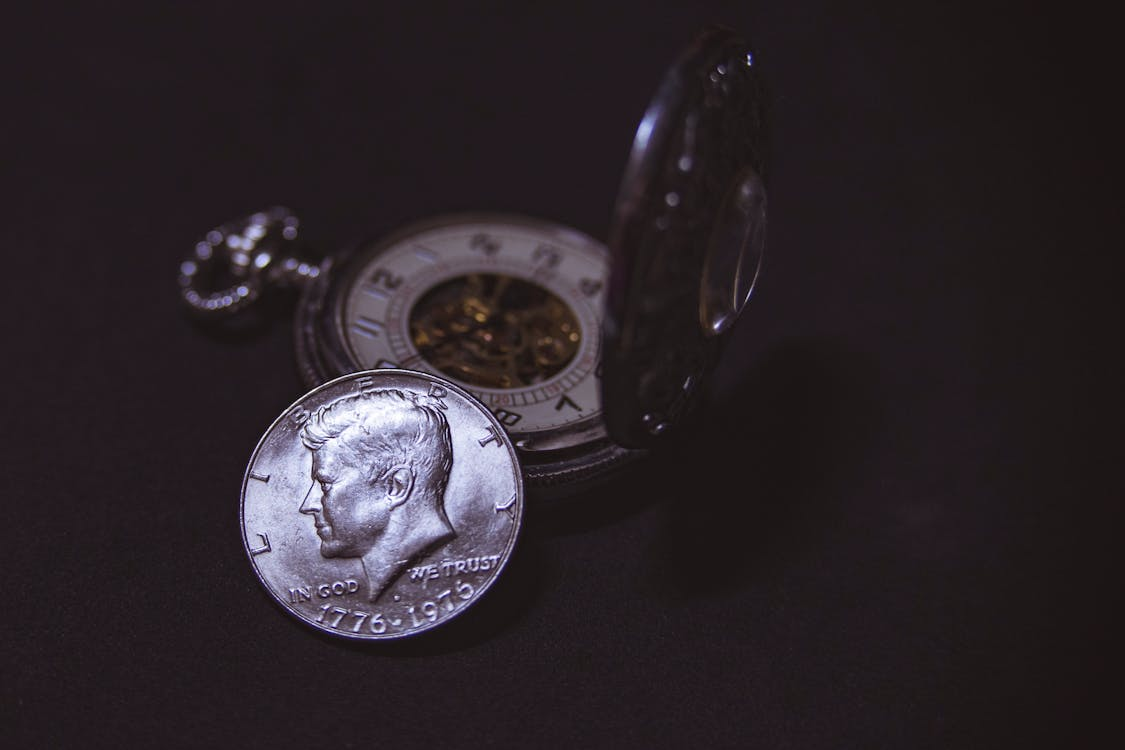 Silver coin with Kennedy’s face and 'In God We Trust' inscription, 1776-1976, backed by a pocket watch on a black background.