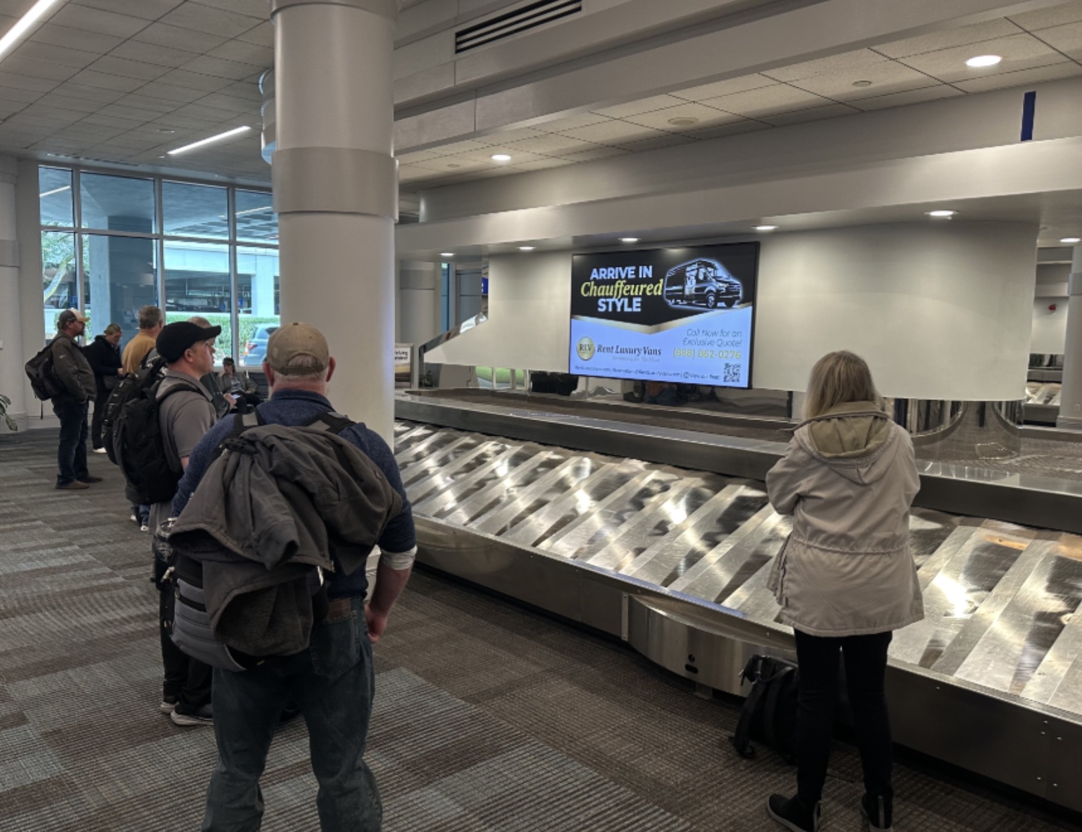 People waiting at baggage claim for their bags at the airport with an TV advertisement for a chauffer company. 
