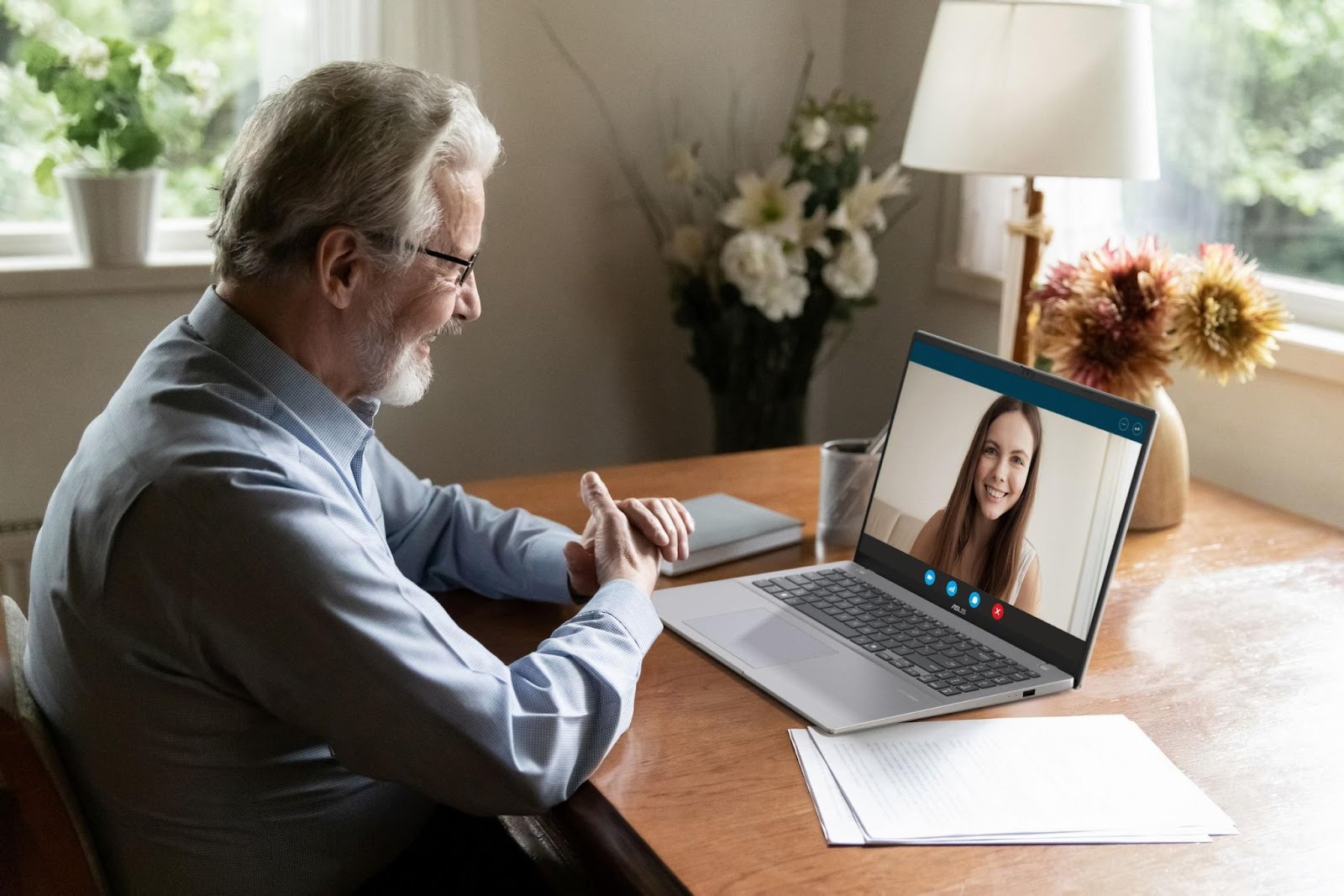 Homem mais velho realizando uma videochamada no ASUS Vivobook 16 em um ambiente doméstico com plantas e flores ao fundo