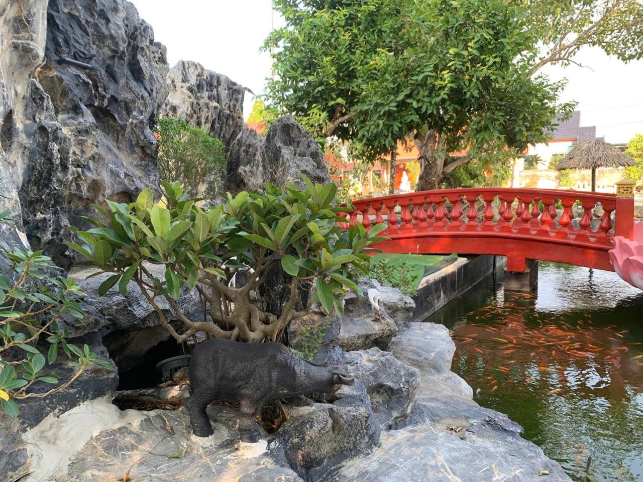 A red bridge over a pond with rocks and a statue of a pigDescription automatically generated