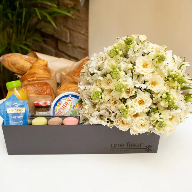 Arranjo de flores brancas e verdes em cesta de café da manhã com pães, sucos, queijos, geléias e macarrons em uma caixa preta
