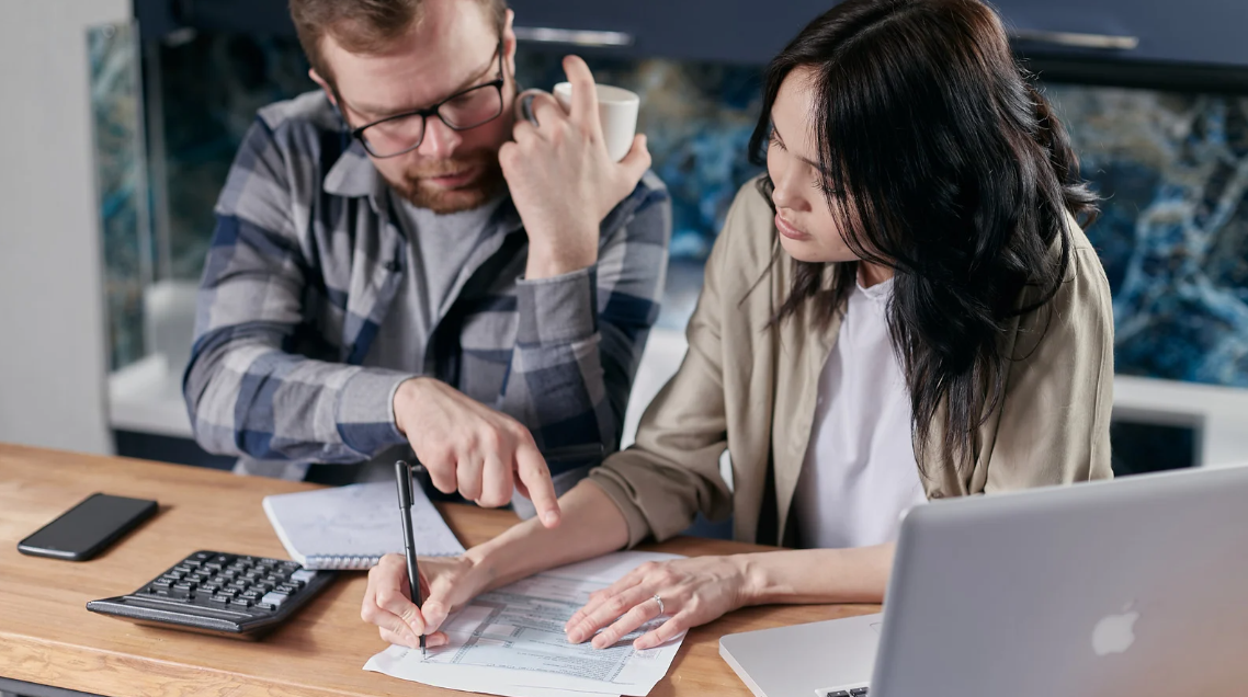 (A couple reviews financial documents together at a kitchen table, using a calculator and laptop to manage bills or taxes.)