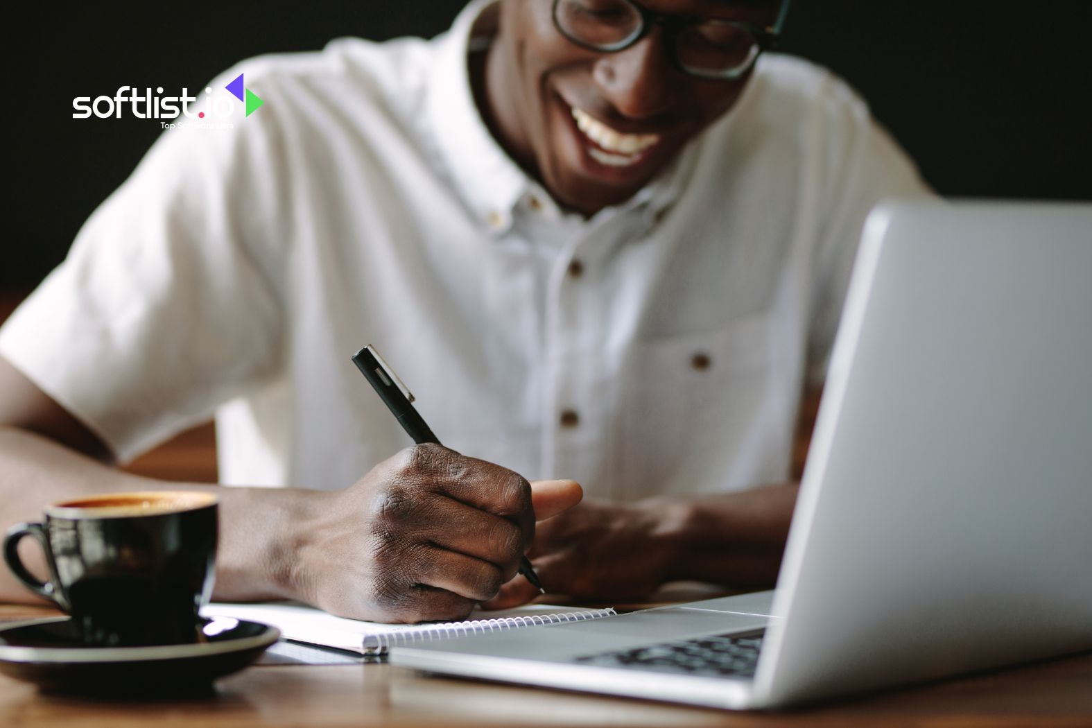 Man smiling while writing in a notebook beside a laptop and coffee cup