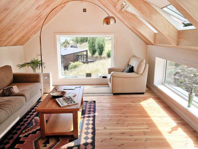 Cozy attic room with wooden floors, a beige armchair, a brown sofa, and a wooden coffee table. Large uPVC windows offer a view of greenery outside, allowing sunlight to illuminate the space.