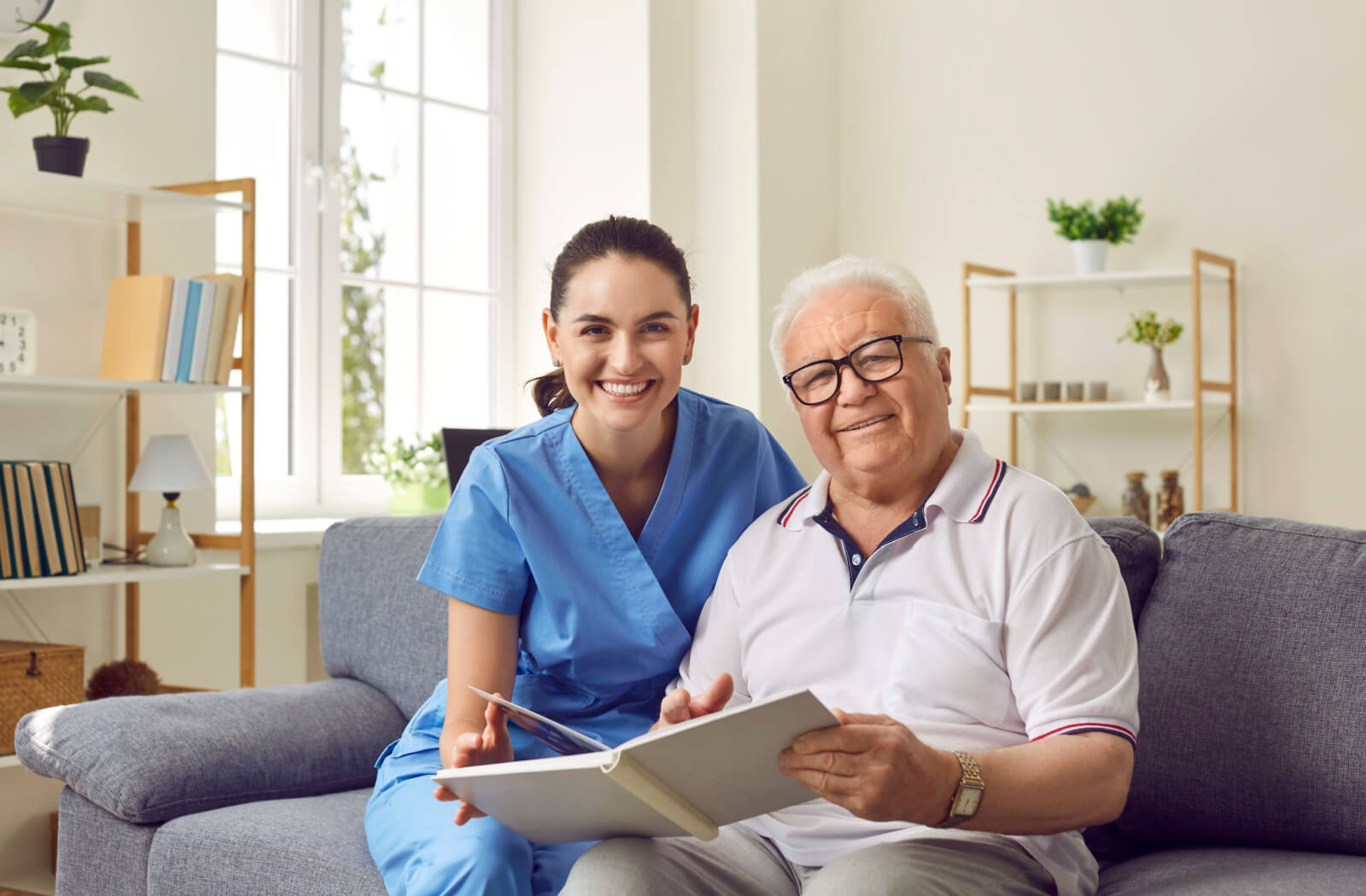 A caregiver and an older adult with dementia in memory care sitting on a couch and smiling while looking through a photo album.