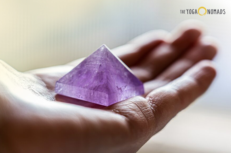 A small, translucent purple pyramid-shaped amethyst crystal resting on the palm of a hand. The background is blurred with soft lighting, highlighting the crystal’s clarity and color.