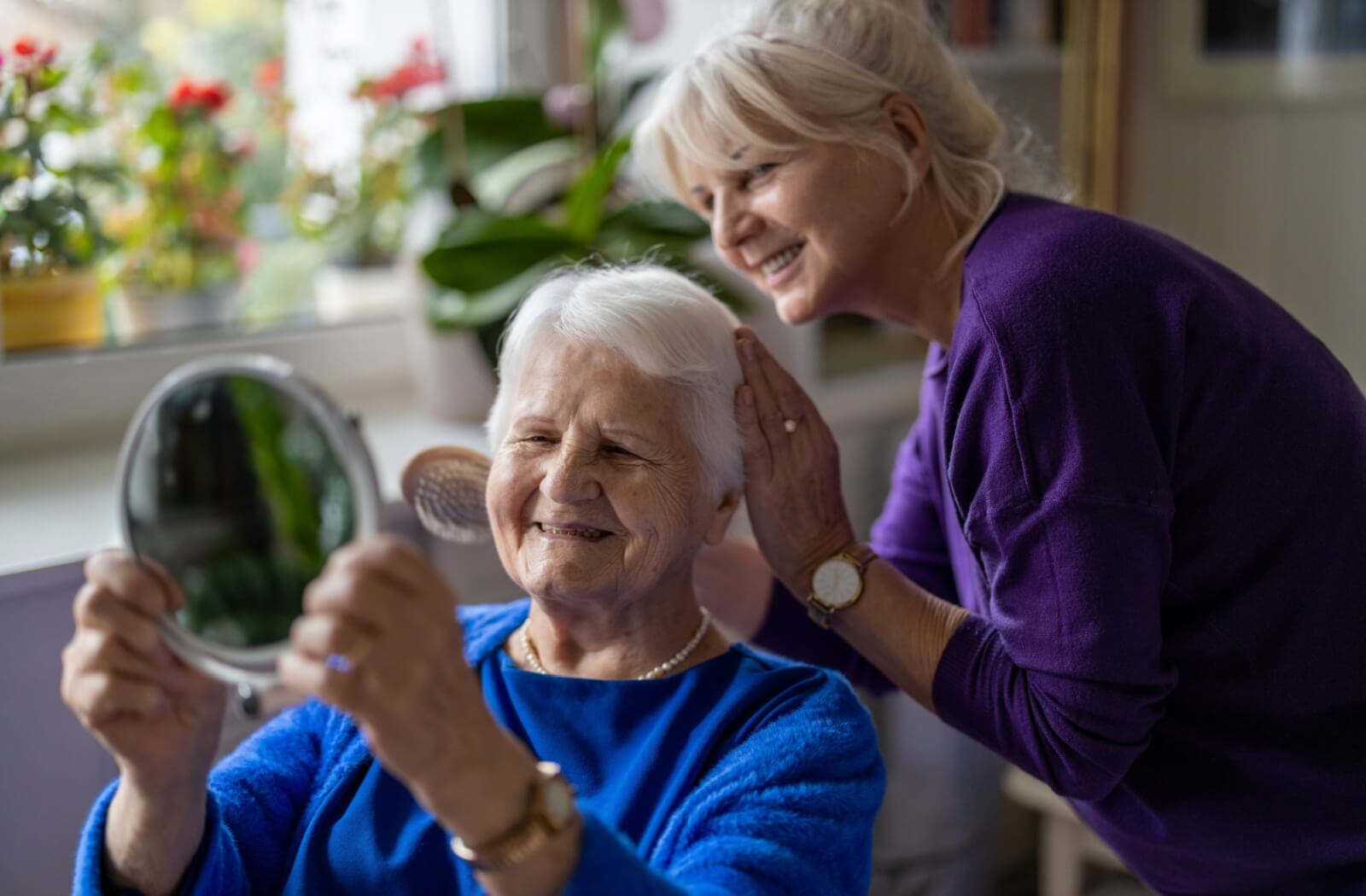 Cheerful elderly woman admiring her reflection in a mirror while her daughter lovingly helps with her hair.