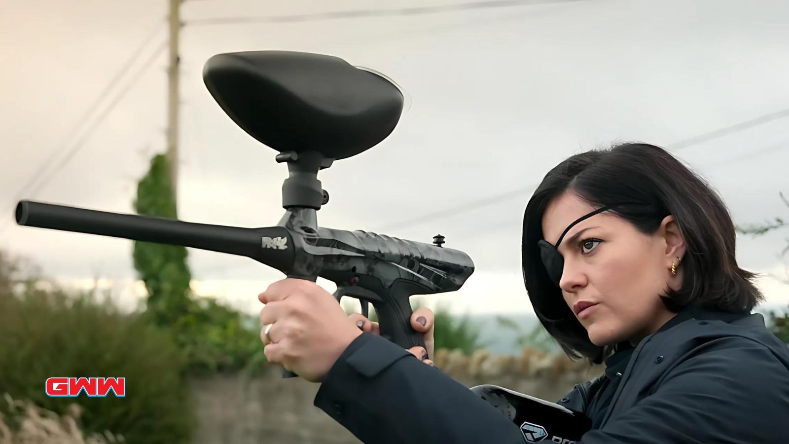 Bibi Garvey with an eyepatch aiming a paintball gun outdoors.