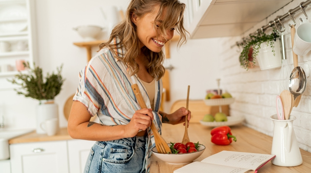 Une femme préparant une salade fraîche dans une cuisine lumineuse, souriante tout en mélangeant des légumes dans un bol avec des ustensiles en bois.