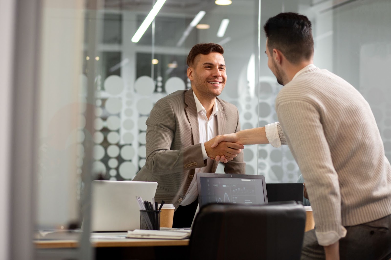 Two professionals shaking hands and smiling in a modern office setting, with laptops, papers, and coffee cups on the desk, indicating a successful meeting or agreement.