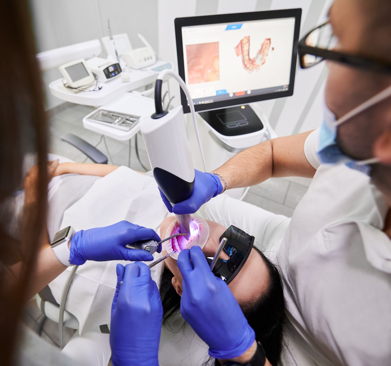 A dental professional uses a modern techniques of intra oral scanner to capture detailed 3D images of a patient's mouth. The patient, wearing protective eyewear, reclines in a dental chair while the dentist and an assistant, both wearing gloves, perform the procedure. A computer screen in the background displays the scanned image of the patient's teeth, emphasizing the use of advanced dental technology in a clinical setting.