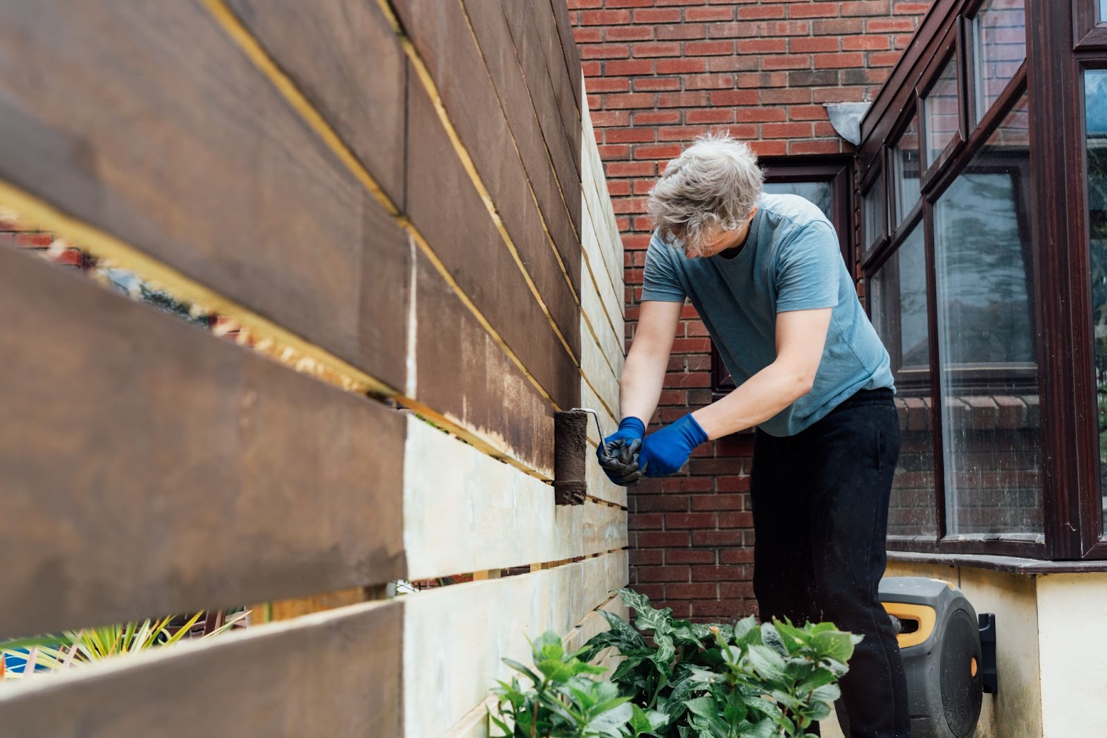 A young man paints a wooden fence with a roller, with the glass walls of a house visible close behind him. 