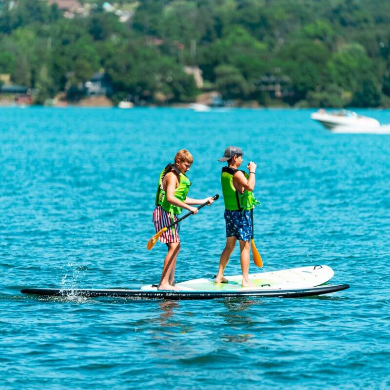 two boys wearing life jackets are stand up paddleboarding on the water at sports camp in FL