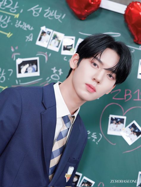 This contains an image of ZEROBASEONE's Yujin in a suit and tie standing next to a chalkboard with pictures on it