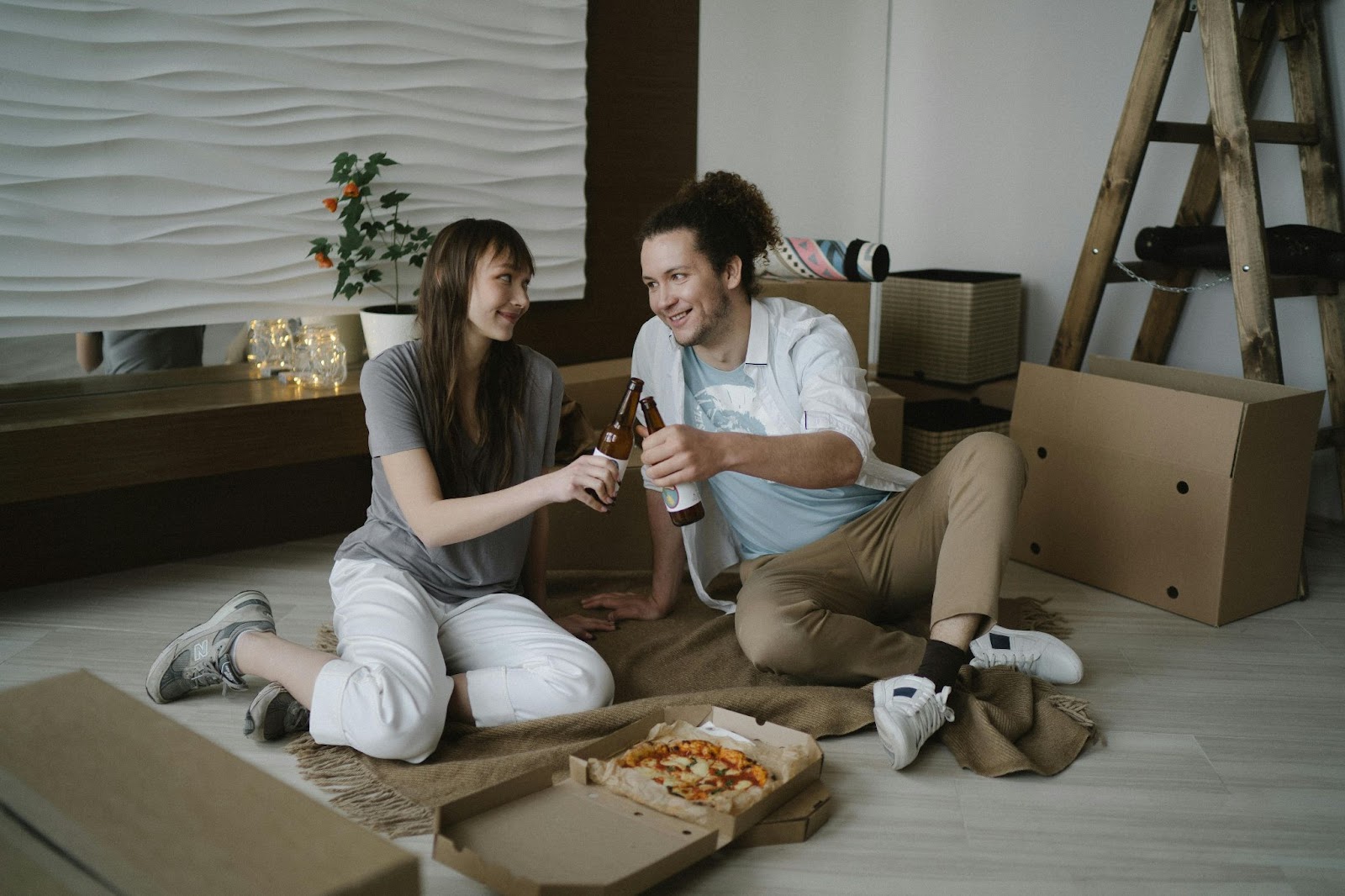 Young couple in Utah sitting on the floor of their new home, toasting with drinks, surrounded by moving boxes and a pizza box, celebrating their move. This is their first home and first mortgage!