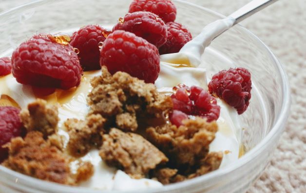 A glass bowl and spoon of healthy weight gain snacks containing Greek yogurt, strawberries, honey, and dry groundnut flakes. 