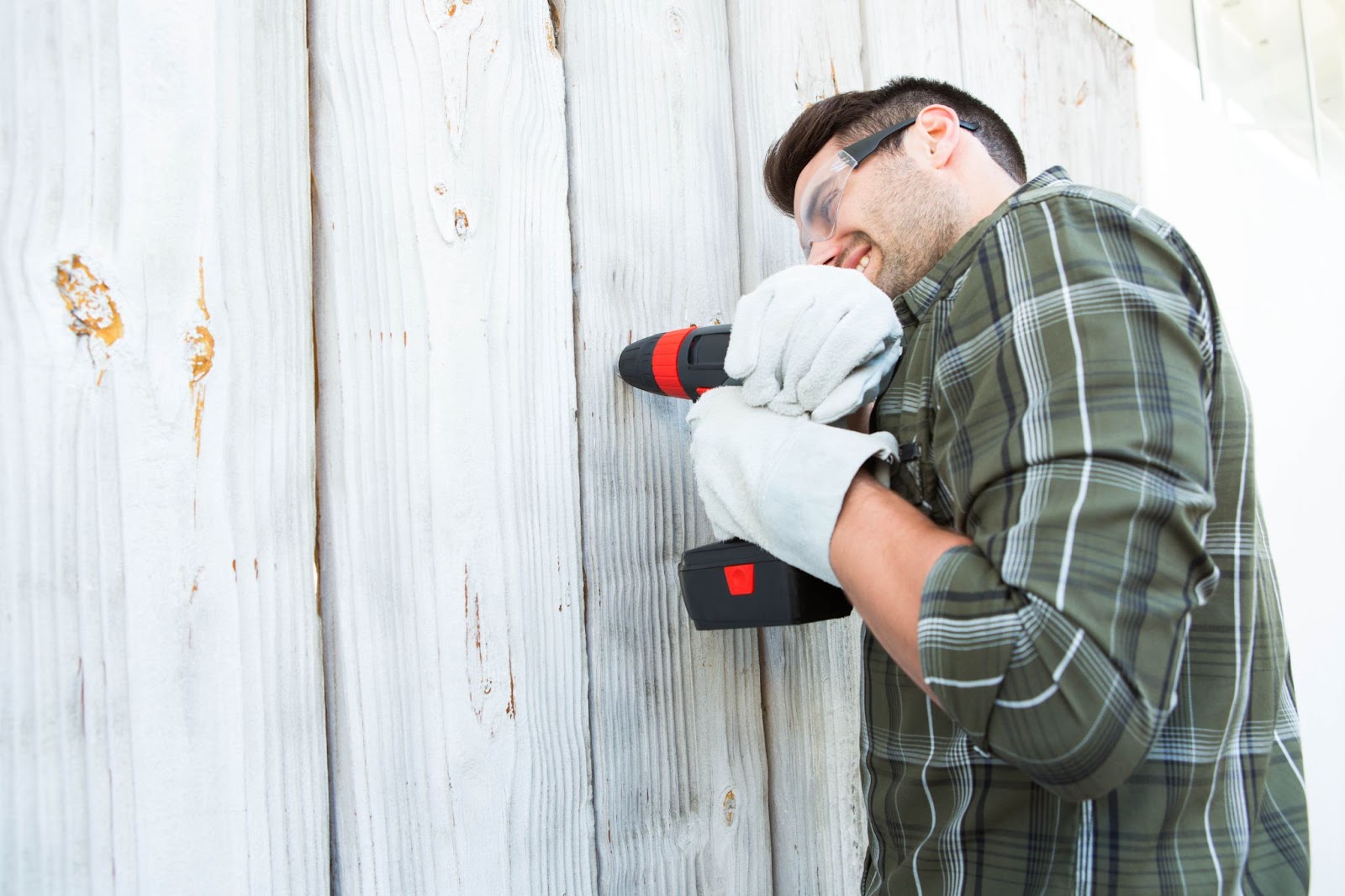 A side view of a construction worker operating a hand drill to secure a wooden fence panel.