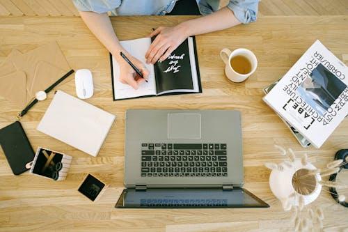 Free A woman writing notes at a home office desk with a laptop and coffee cup. Stock Photo