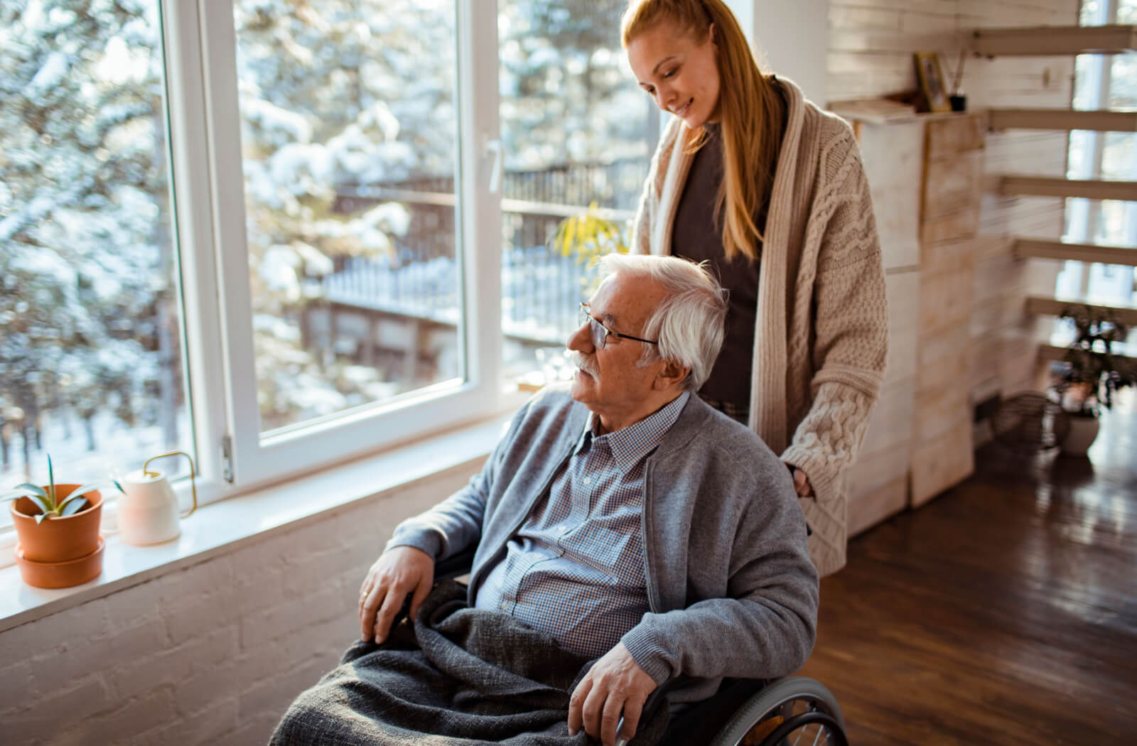 A grandchild helping their grandparent while looking at the snow outside through a window in senior living.