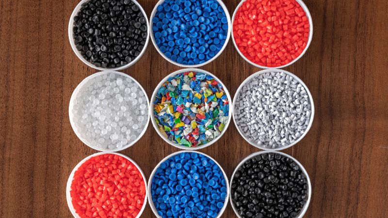 Nine bowls with varied colored plastic pellets—black, blue, red, white, and multicolored—arranged on a wooden surface.