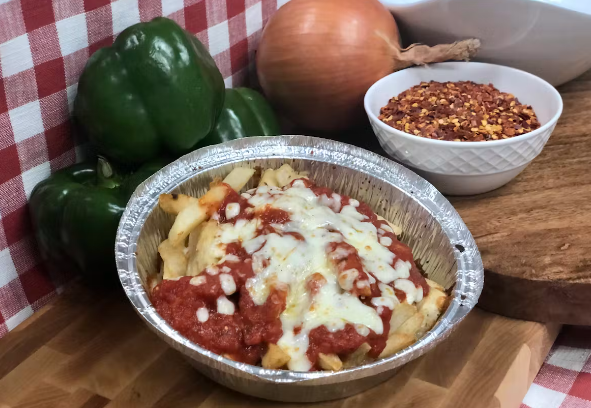 Fries in a cup seasoned with pizza sauce and cheese placed on a wooden board, with capsicums, chilli flakes and an onion placed behind it.