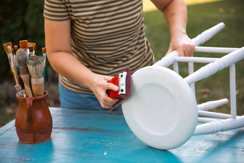 A person is sanding a white wooden chair outdoors, preparing it for a fresh coat of paint. Nearby, a jar filled with paintbrushes sits on a blue table, emphasizing the DIY nature of the outdoor furniture project.