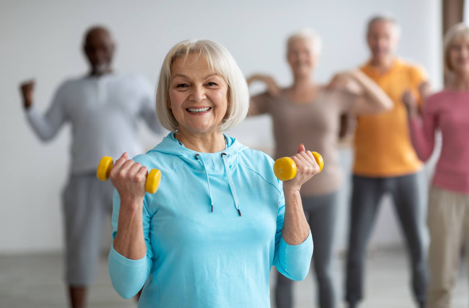 A smiling older adult during a fitness class holding two small yellow weights.