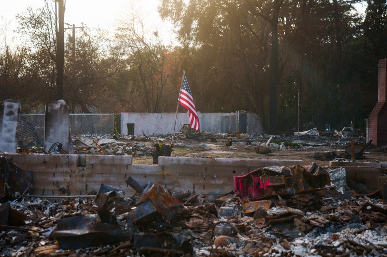 A tattered American flag stands amid the ruins of a burned-down structure, surrounded by charred debris and remnants of walls. The ground is covered in ash and broken materials. In the background, leafless trees and a partially collapsed fence add to the scene of destruction. The golden light of the setting or rising sun filters through the trees, casting a somber glow over the aftermath.