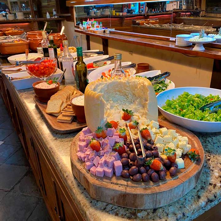 Buffet de saladas e queijos com diversos ingredientes frescos, incluindo folhas verdes, tomates, queijo, presunto, frutas e azeite, disponível para café da manhã ou almoço.