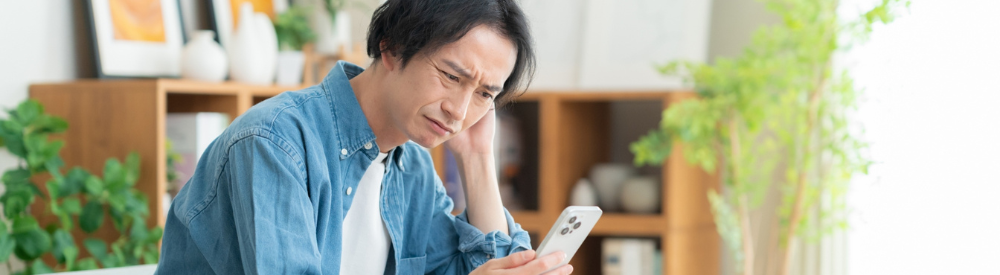 自宅でスマートフォンを見ながら、困った表情で頭を抱える男性