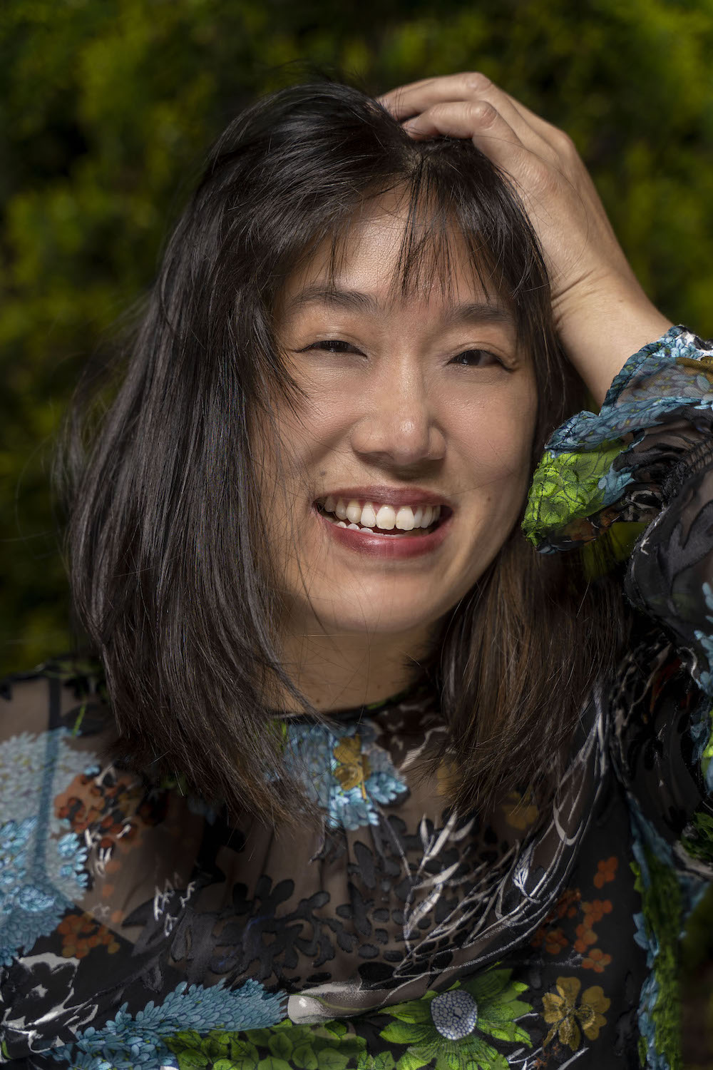 A smiling woman with long straight shoulder-length black hair and brown eyes, wearing a green blue and black floral top, with her left hand resting on her head.