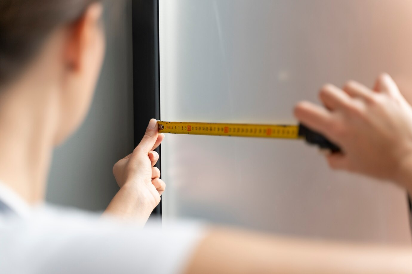 A person measuring the width of a Garage Door or reflective surface with a yellow tape measure, focusing on accuracy and alignment, with natural light illuminating the workspace