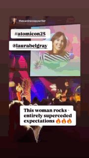 A GIF shows a speaker on stage at Atomicon, with a large screen behind her displaying an image of Laura Belgray smiling against a colorful background. The stage is lit with vibrant pink, purple, and blue lighting, and a live band plays beside her. Text overlays include “#atomicon25,” “@laurabelgray,” and a caption that reads, “This woman rocks – entirely superceded expectations” followed by three fire emojis. The audience and energetic atmosphere suggest a lively, engaging event.