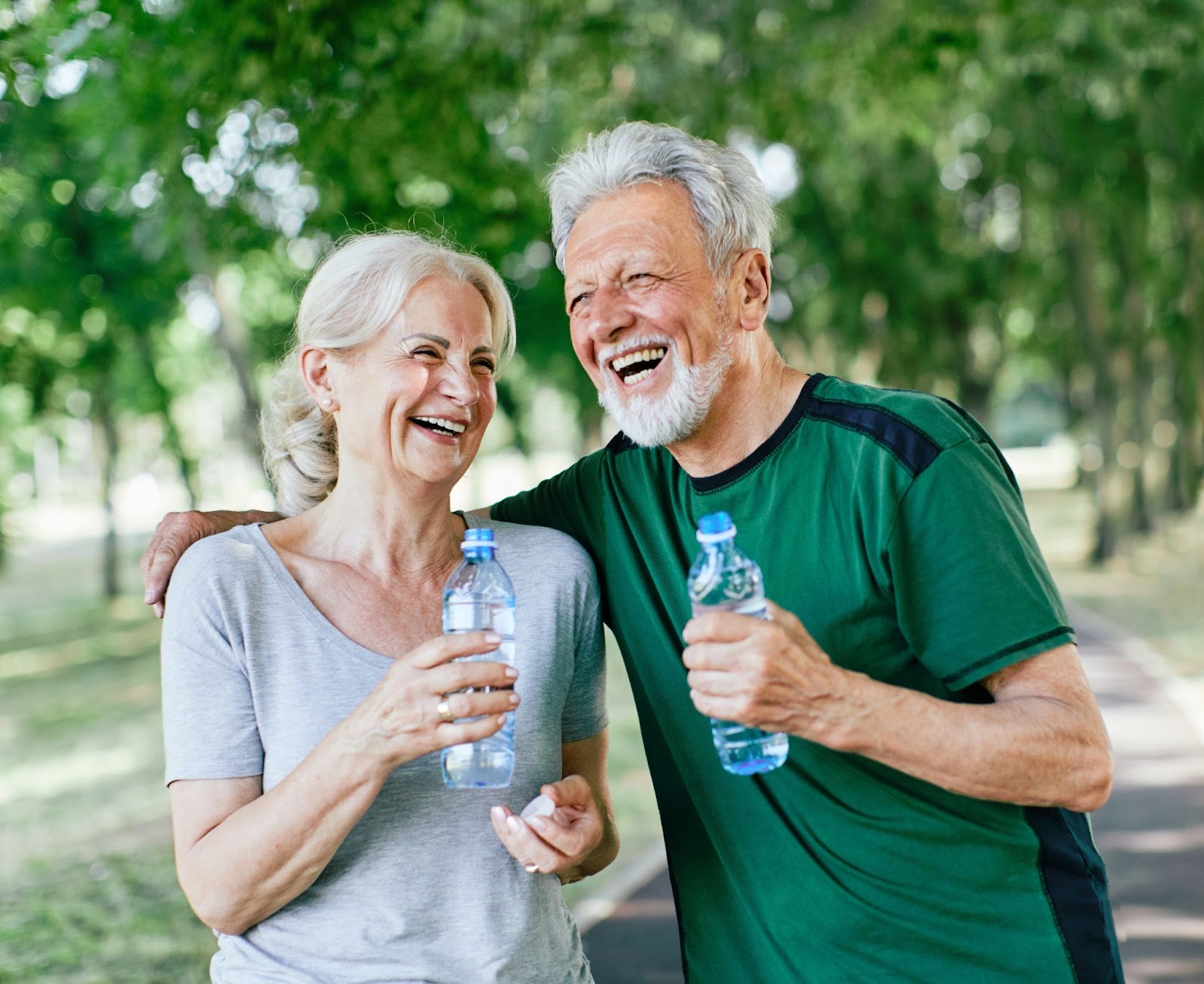 An elderly couple laugh with water bottles in their hands after walking outdoors.