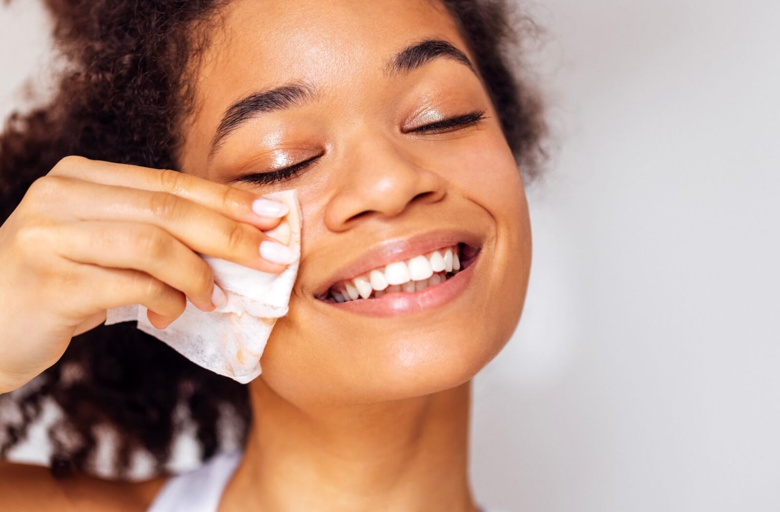 A woman smiles as she uses a gentle makeup wipe to remove makeup from her cheek and lower eyelid