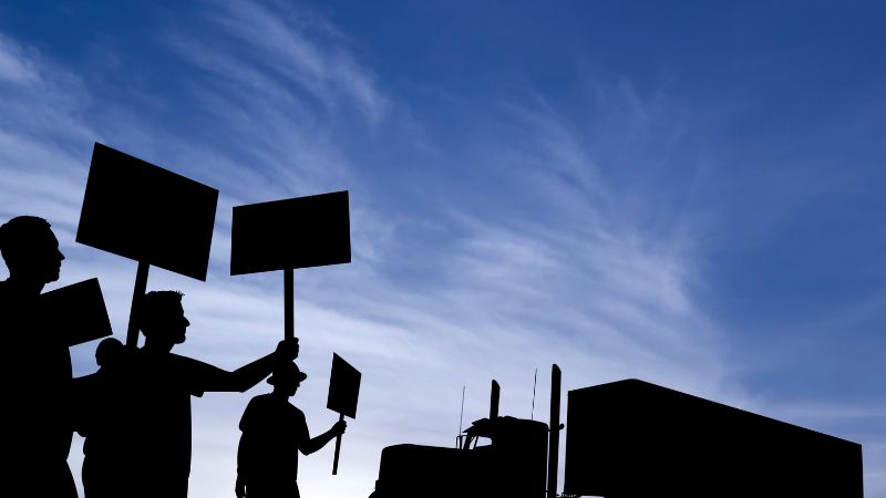 Des silhouettes portant des pancartes de protestation se détachent devant un gros camion, sur fond de ciel bleu.