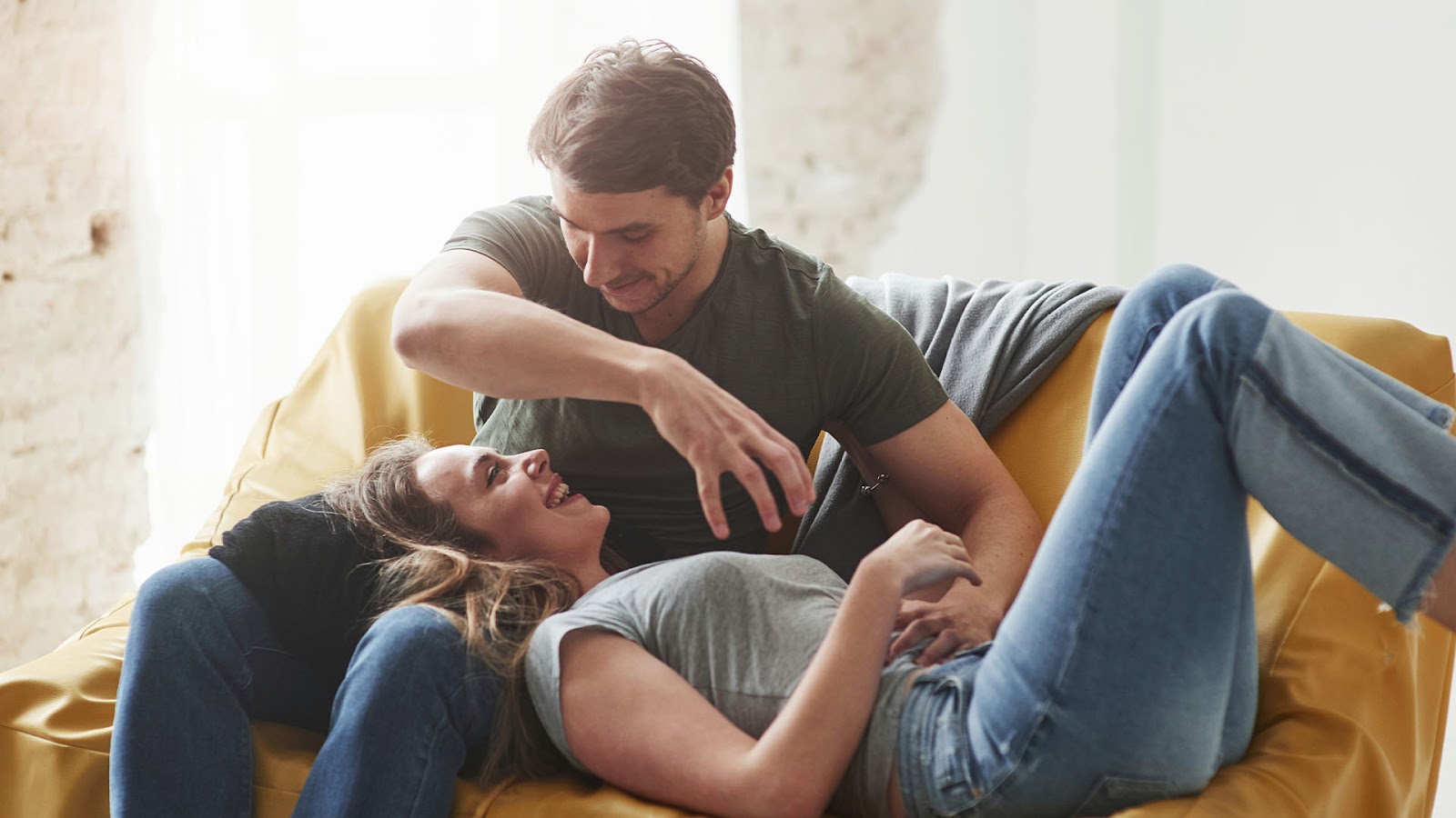 Brunette man and woman, the man is sitting on a yellow couch while the woman rests her head on his lap