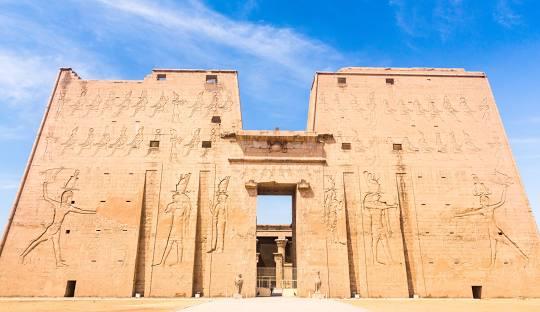 A large stone building with carvings on it with Temple of Edfu in the background

Description automatically generated