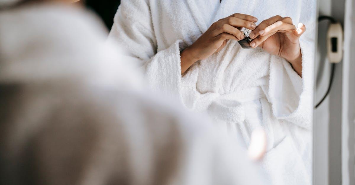 Crop unrecognizable young lady in white bathrobe opening face cream while standing in front of mirror after taking shower in morning