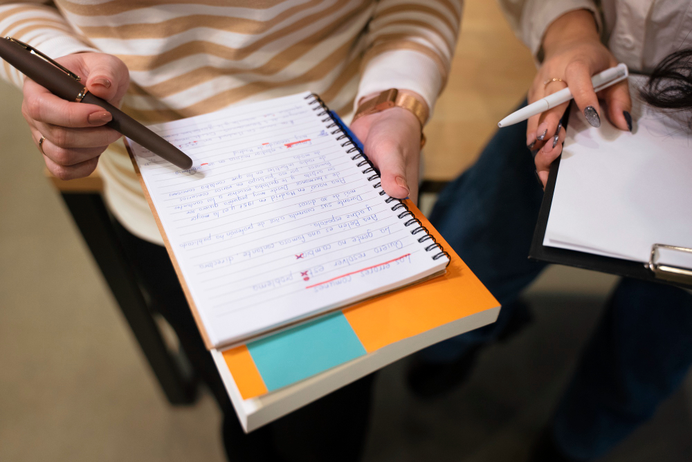 Two people are closely examining notes. One holds a spiral notebook with handwritten text and corrections, while the other clutches a clipboard and pen. They seem to be exchanging study tips for an upcoming GATE Architecture 2024 exam.