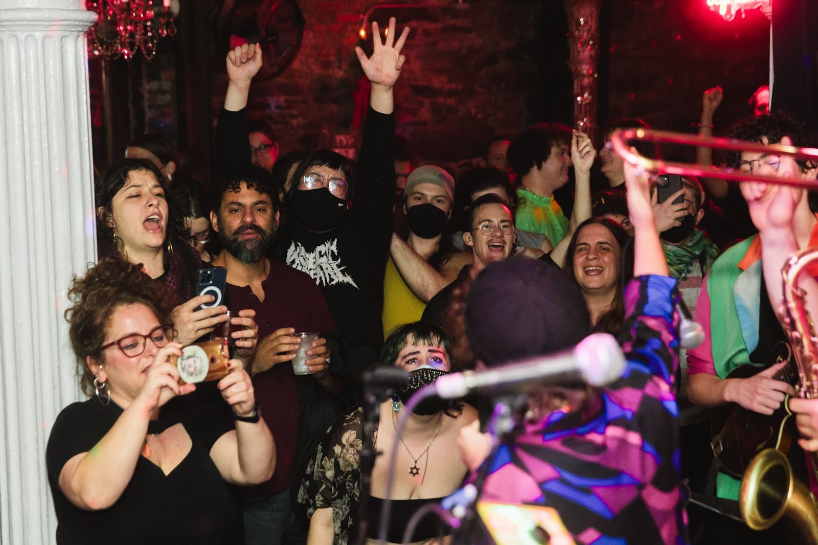 A crowd of people, some with hands raised, some masked, and one up front wearing a Magen David necklace, enjoying a Chaia performance.