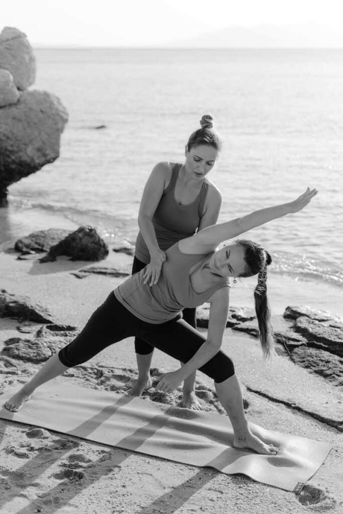 Two individuals are on a beach, with one person assisting the other in performing a yoga pose. The person being assisted is in a side angle pose, with one arm extended overhead and the other arm resting on their bent knee. The assistant is providing support by holding the person's waist and adjusting their posture. The scene is set against a backdrop of the sea and rocks, creating a serene and natural environment for the yoga practice.