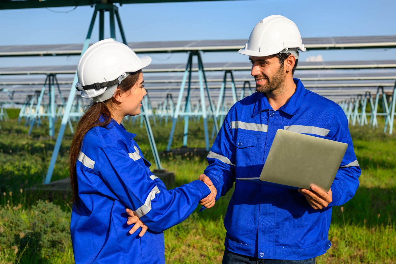 Two field service technicians in blue coveralls and white helmets shake hands after successfully installing solar panels at a solar farm, ensuring efficient energy production.