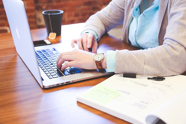 A person typing on a laptop with a cup of coffee and papers on a desk.