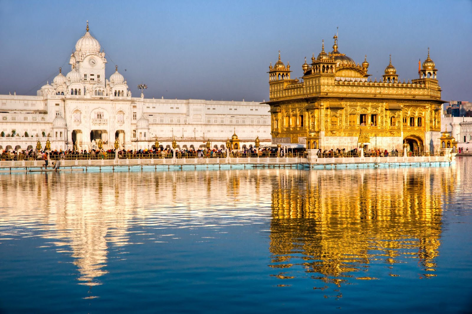 A golden temple with a white building in the background with Harmandir Sahib in the background

Description automatically generated