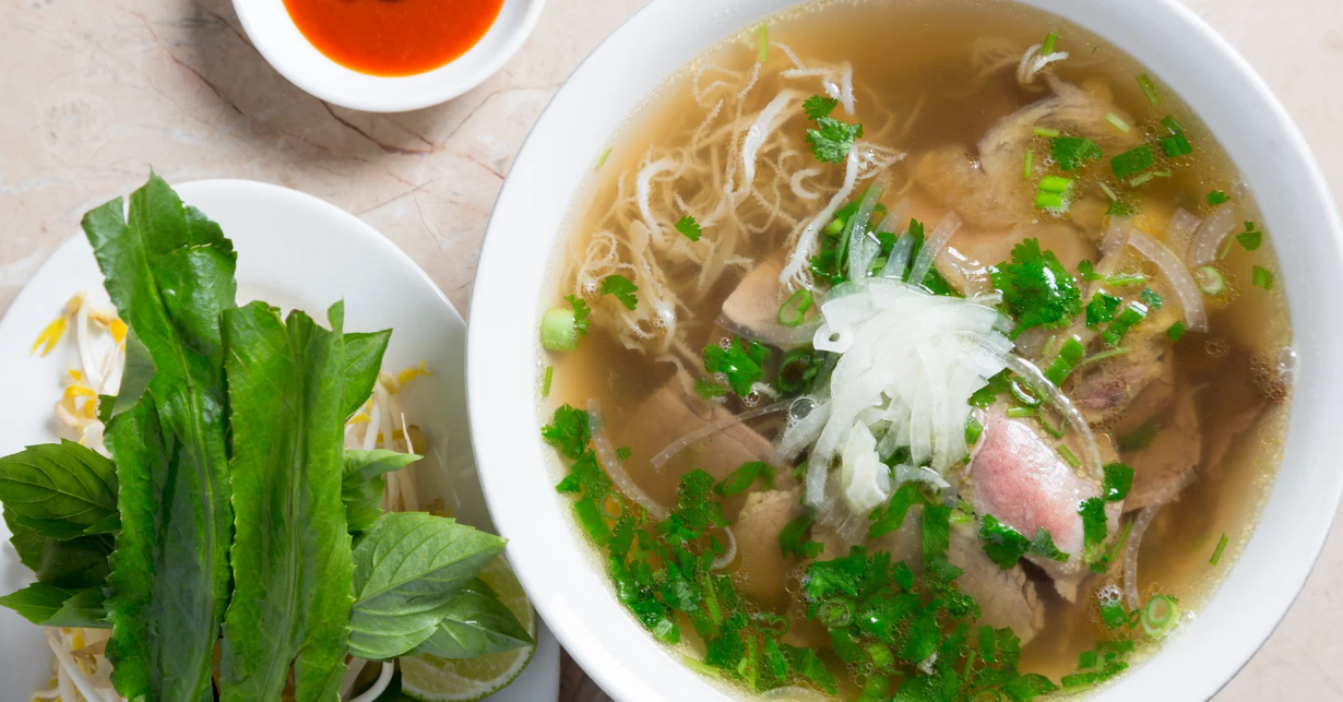 (Eine klassische Schüssel Pho mit klarer, reichhaltiger Brühe und zartem Rindfleisch – serviert mit einem Teller frischer Kräuter, Sprossen und Limette für die perfekte traditionelle Note.)