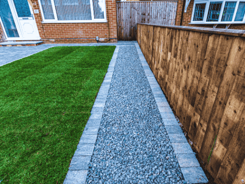 Artificial grass front yard idea with a stone-edged gravel pathway alongside a wooden fence and brick house facade.